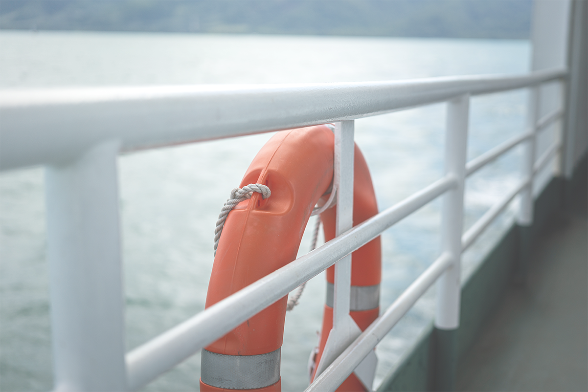 An einem weißen Geländer ist ein orangener Rettungsring befestigt; im Hintergrund sieht man das Meer.