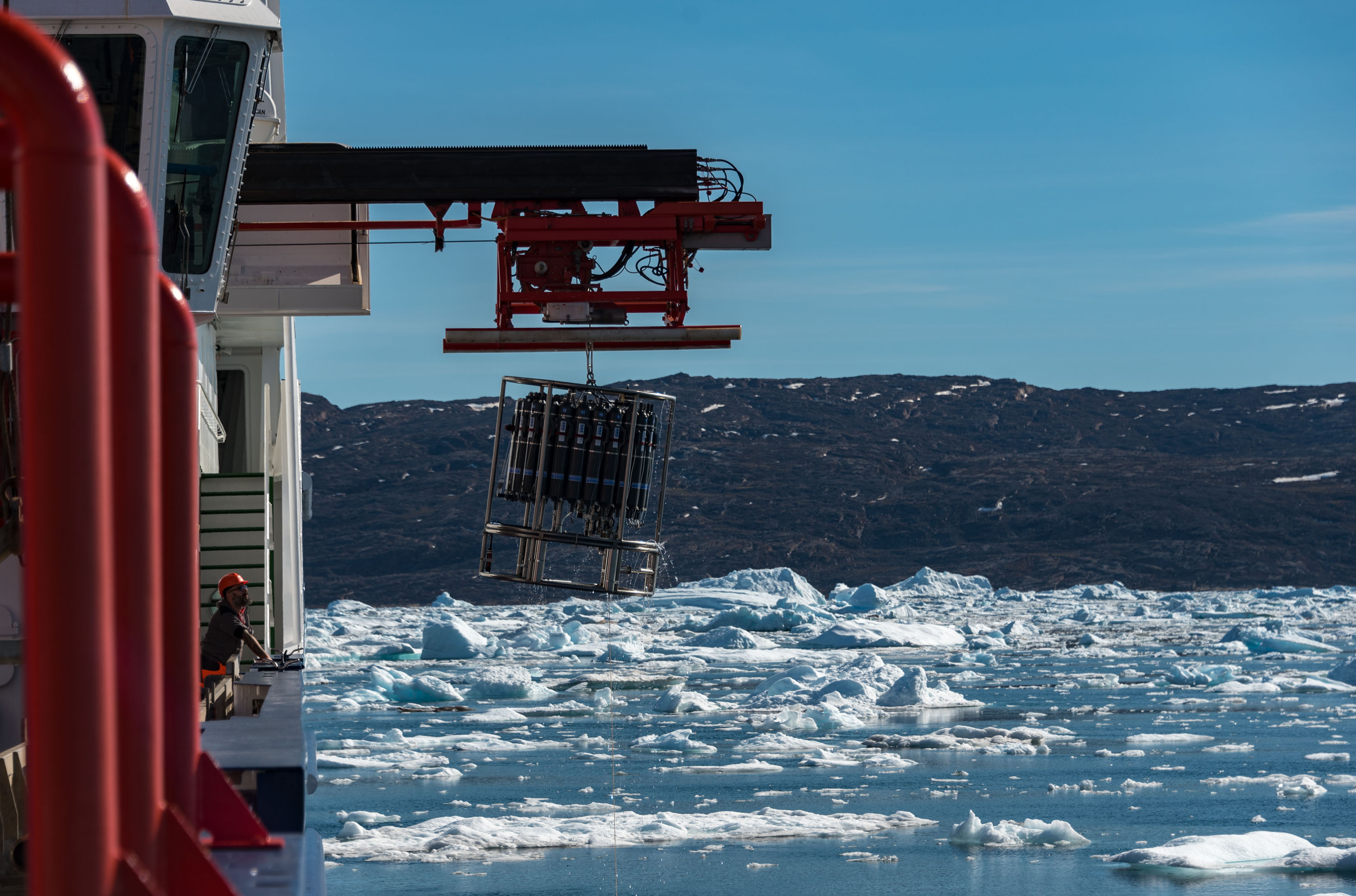 A member of staff is lowering the CTD into the sea. Snow-capped mountains can be seen in the background.